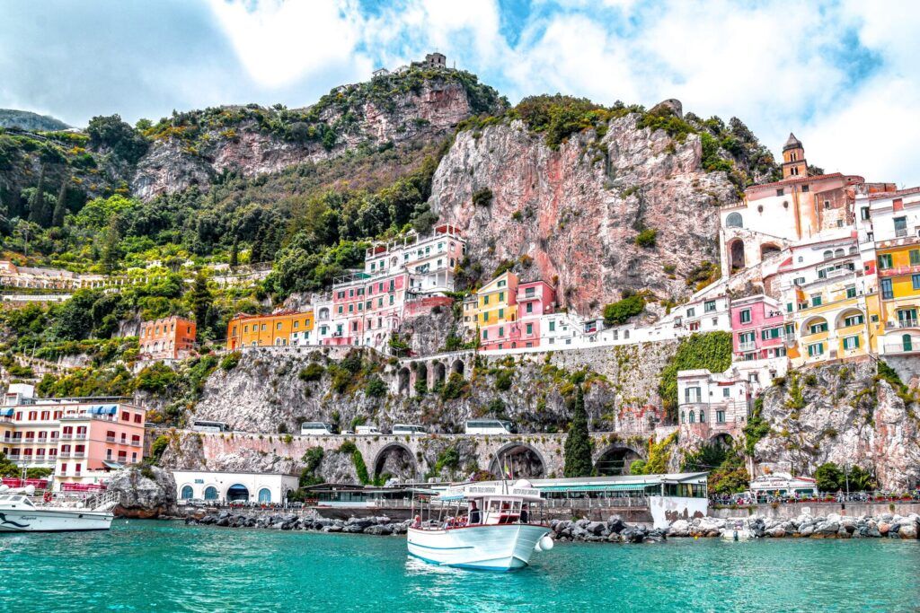 boat in the middle of the sea with colourful houses behind it on a high, rocky cliff