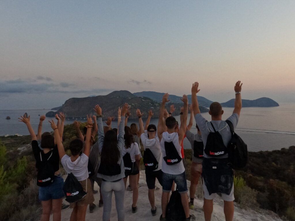 A group of WeRoad travelers cheering with arms raised while looking at the sea and islands at sunset.