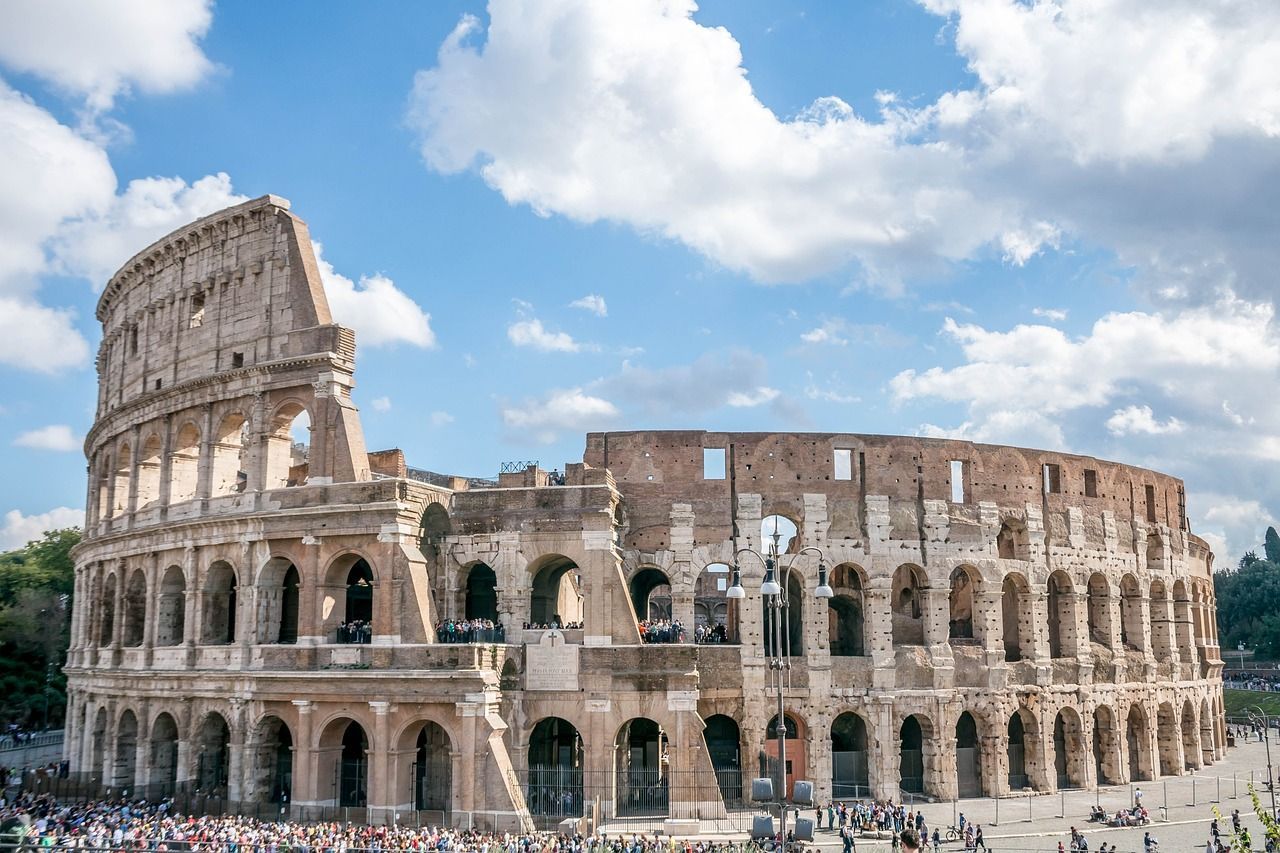The Colosseum in Rome, seen on a sunny day with clouds above, filled with tourists around its perimeter
