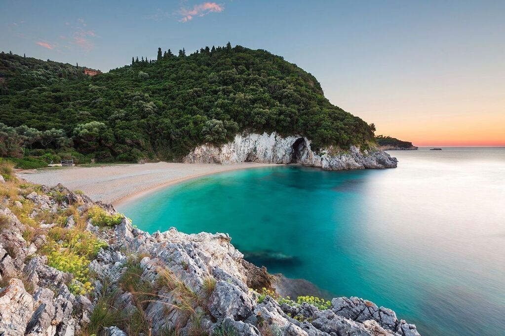 Secluded beach with crystal-clear turquoise water, surrounded by rocky cliffs and lush green hills at sunset in Corfù