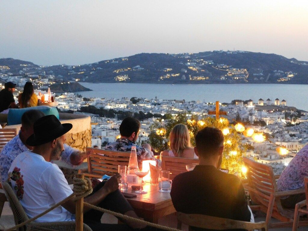 People enjoying a candlelit dinner at a rooftop restaurant in Mykonos, overlooking the illuminated town and sea at dusk