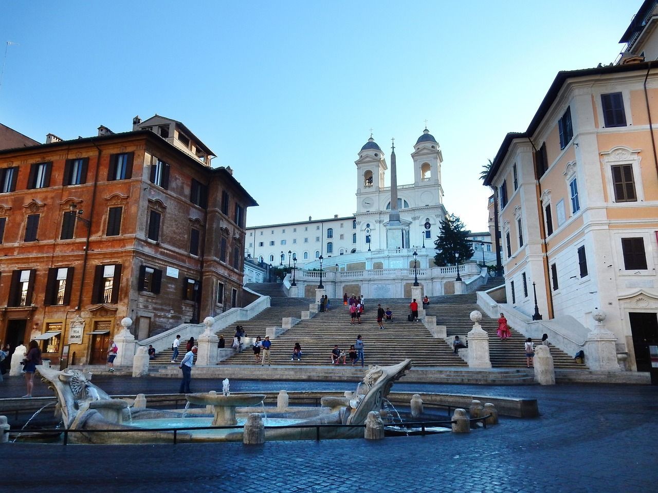 The Spanish Steps in Rome, bustling with tourists, leading up to the Trinità dei Monti church at the top, under a clear blue sky.