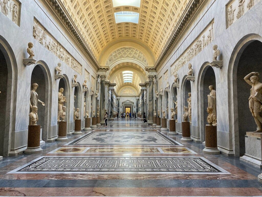 Long corridor inside the Vatican Museums, flanked by classical statues and leading to a beautifully adorned ceiling