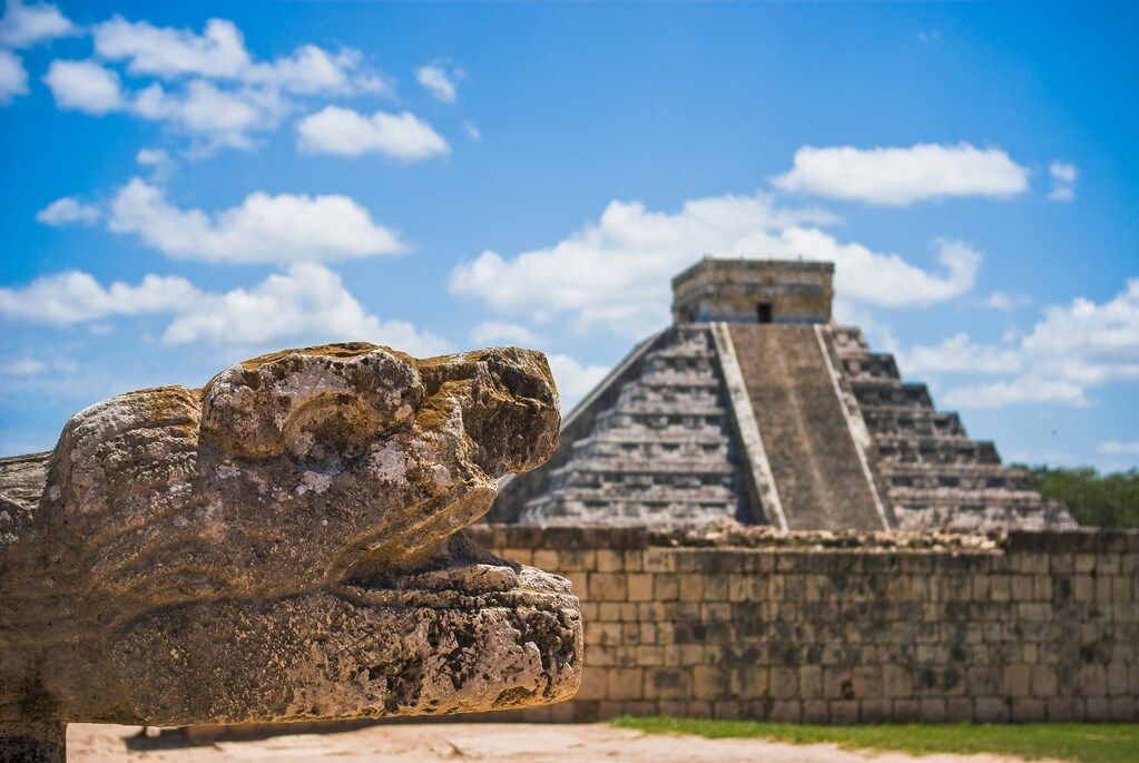 Close-up of a stone serpent head sculpture at Chichén Itzá, with the iconic Temple of Kukulcán pyramid