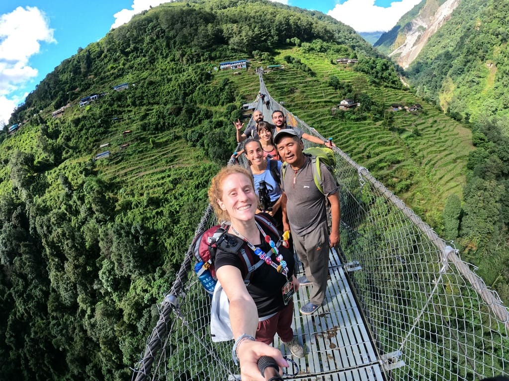 A group photo during a WeRoad group trip on a suspension bridge surrounded by lush green hills in Nepal