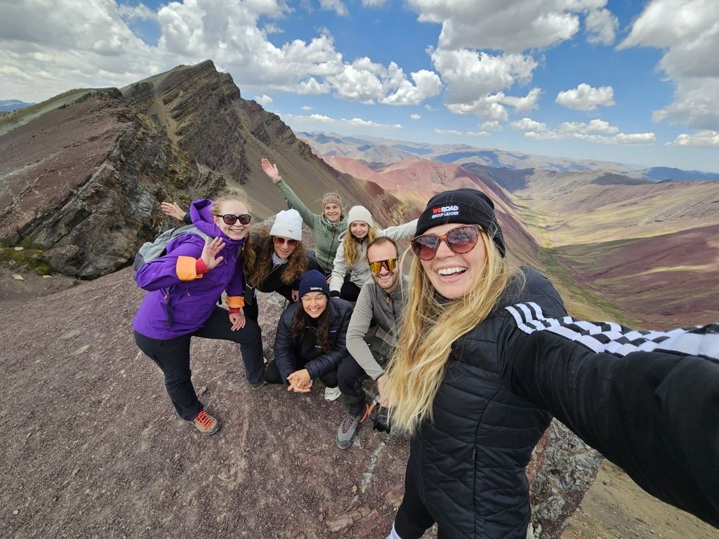 Group of smiling hikers on a mountain trail in Peru with colorful peaks in the background during a WeRoad trip.