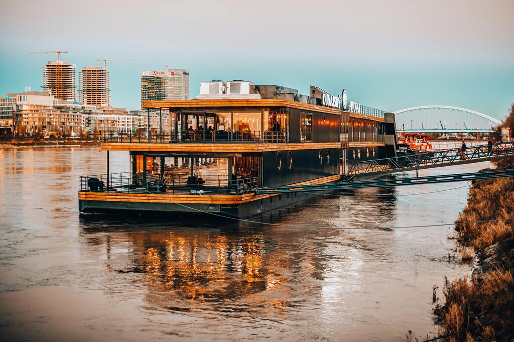 A modern floating restaurant on the Danube River in Bratislava during sunset, reflecting on the water.