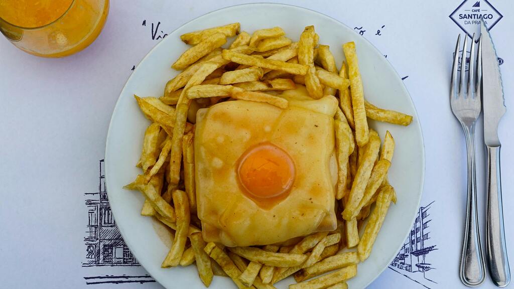 A plate of "Francesinha," a Portuguese sandwich topped with melted cheese, egg, and served with fries and sauce.