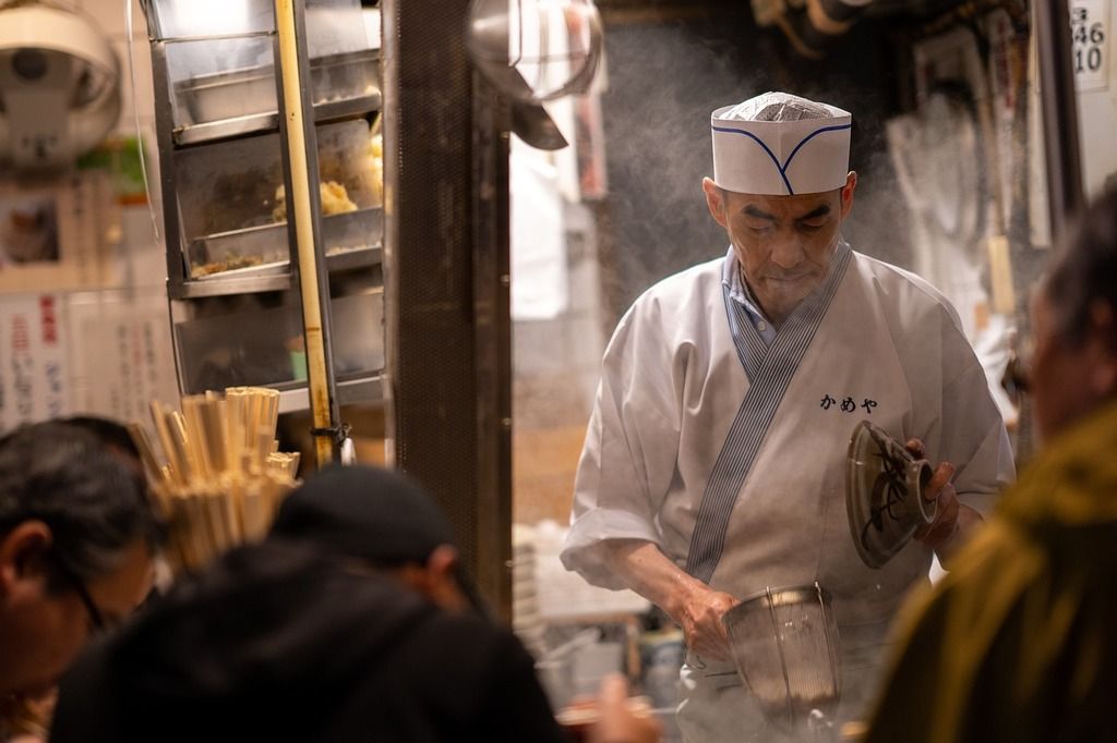 A Japanese chef preparing a dish in a traditional ramen shop, surrounded by a lively kitchen setting.