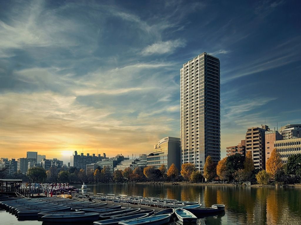 Sunset view of a modern high-rise building overlooking a peaceful lake with small boats docked, surrounded by autumn-colored trees in Tokyo, Japan.