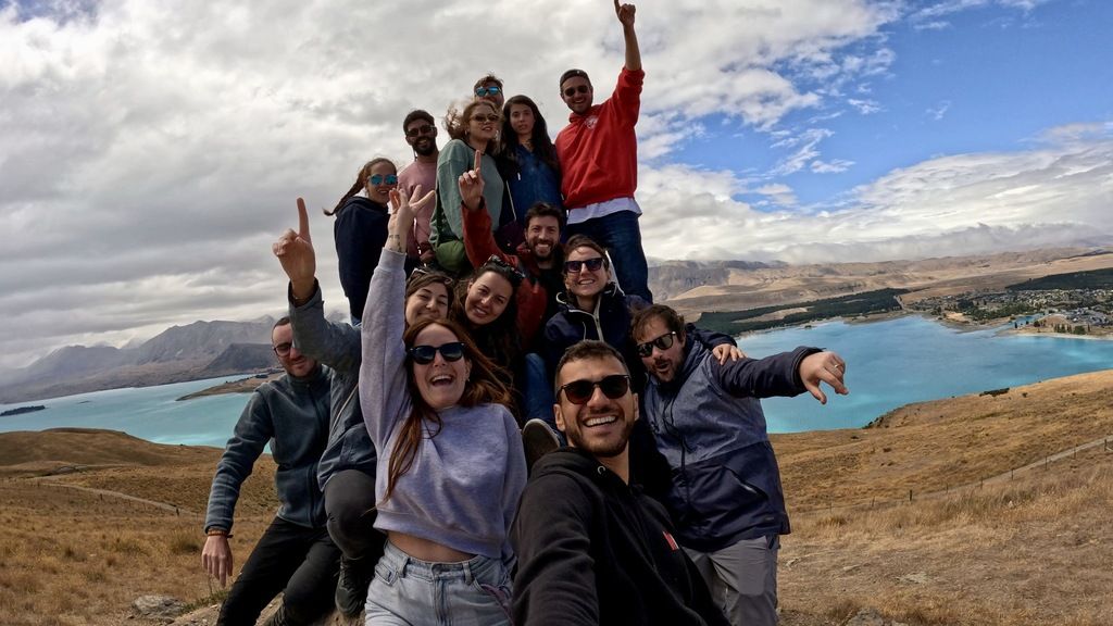 WeRoad group trip in New Zealand with smiling travelers posing on a hill overlooking Lake Tekapo's turquoise waters and surrounding mountains.