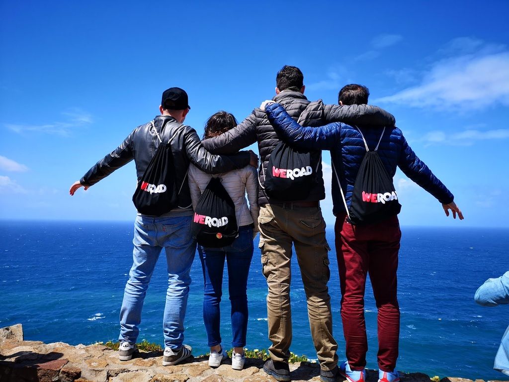 Group of travelers with WeRoad backpacks overlooking the ocean, part of a group trip in Portugal.