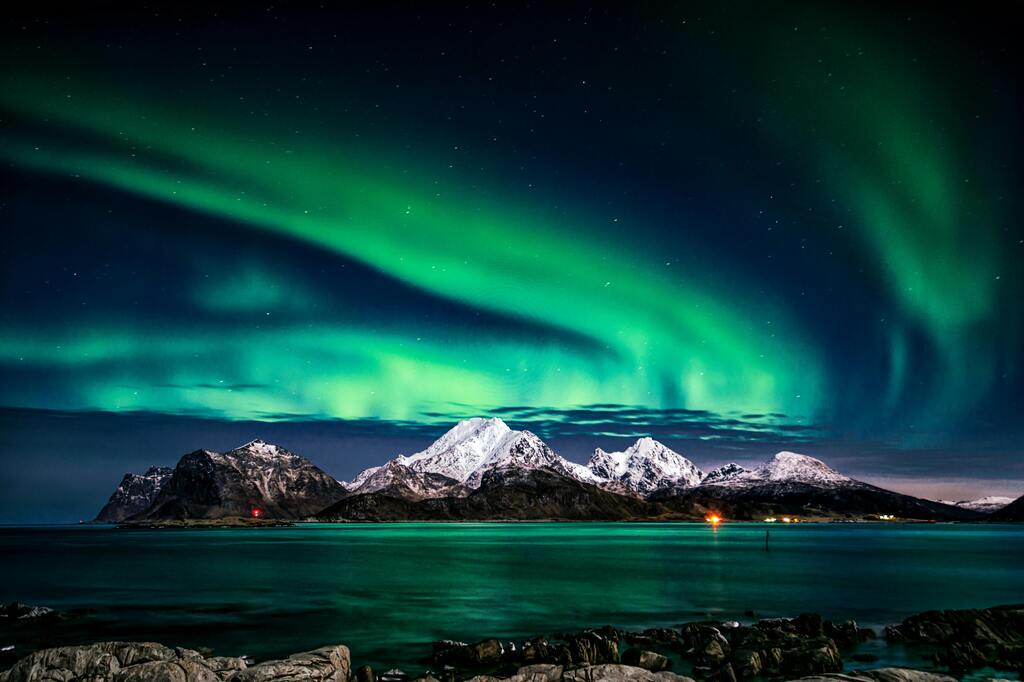 Vivid green northern lights dancing above snow-capped mountains and a serene fjord in Norway.