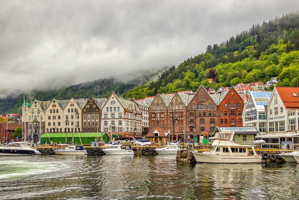 Colorful wooden buildings along the Bryggen waterfront in Bergen, Norway, with boats and a misty forest in the background.