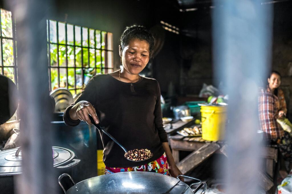 A Cambodian woman cooking in a rustic kitchen, stirring a wok over an open flame.
