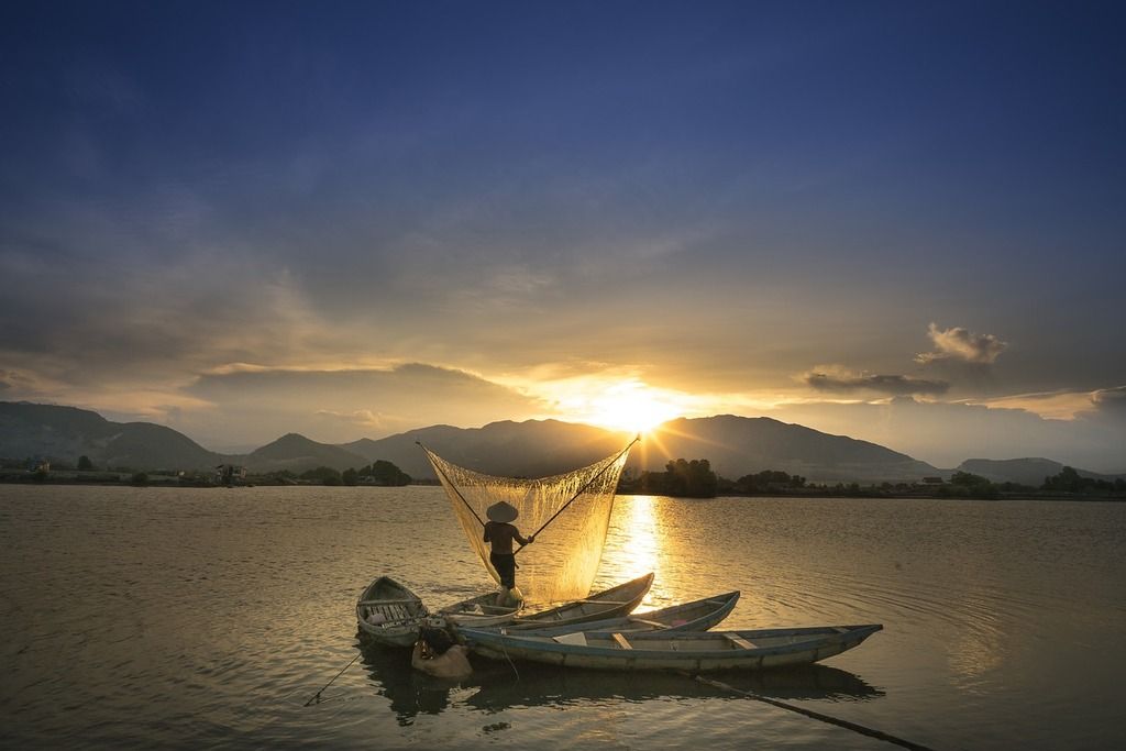 A fisherman casting his net at sunset on a peaceful river, with mountains in the background.
