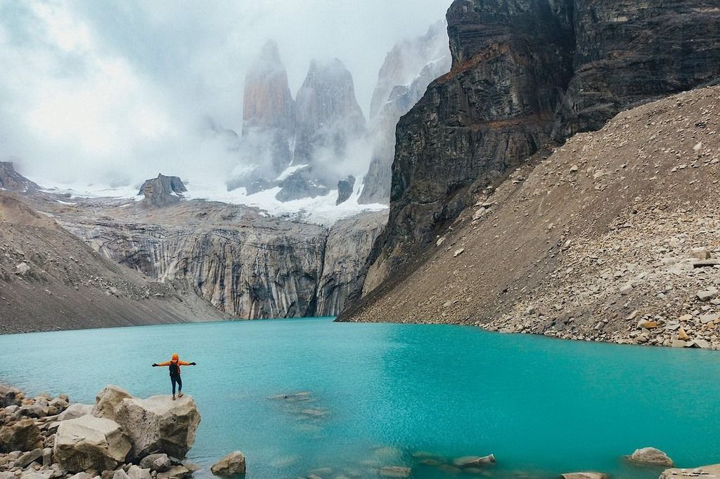 A traveler in an orange jacket stands on a rock overlooking the turquoise waters of Torres del Paine National Park, surrounded by towering granite peaks and misty clouds.