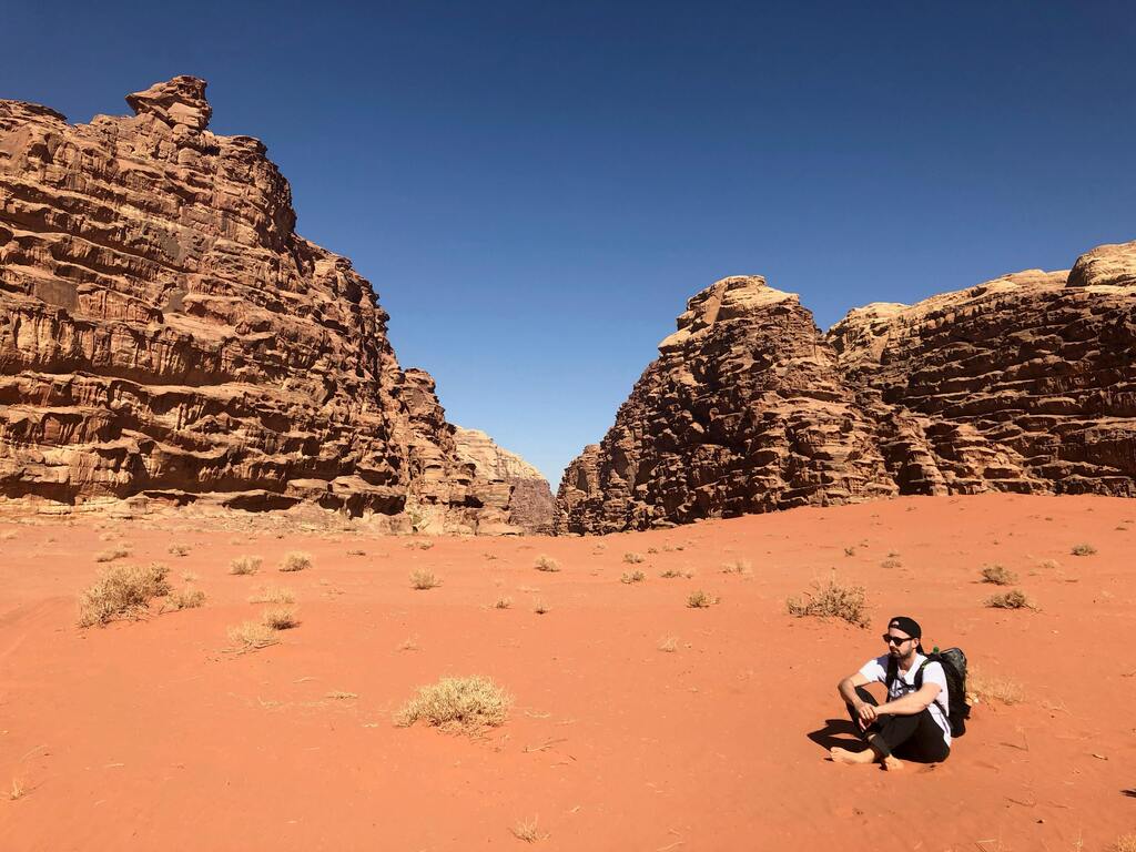 A traveler sitting on the red sand in Wadi Rum, surrounded by towering rock formations under a deep blue sky.