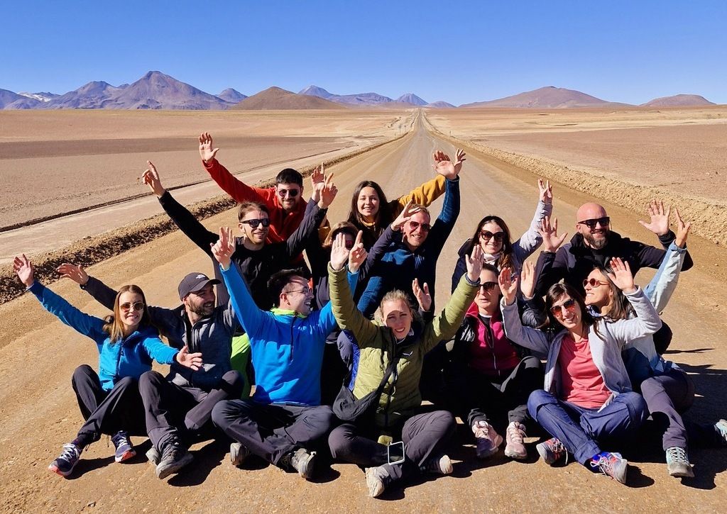 A group of travelers posing on a long, empty desert road in the Atacama Desert, with mountains in the background.