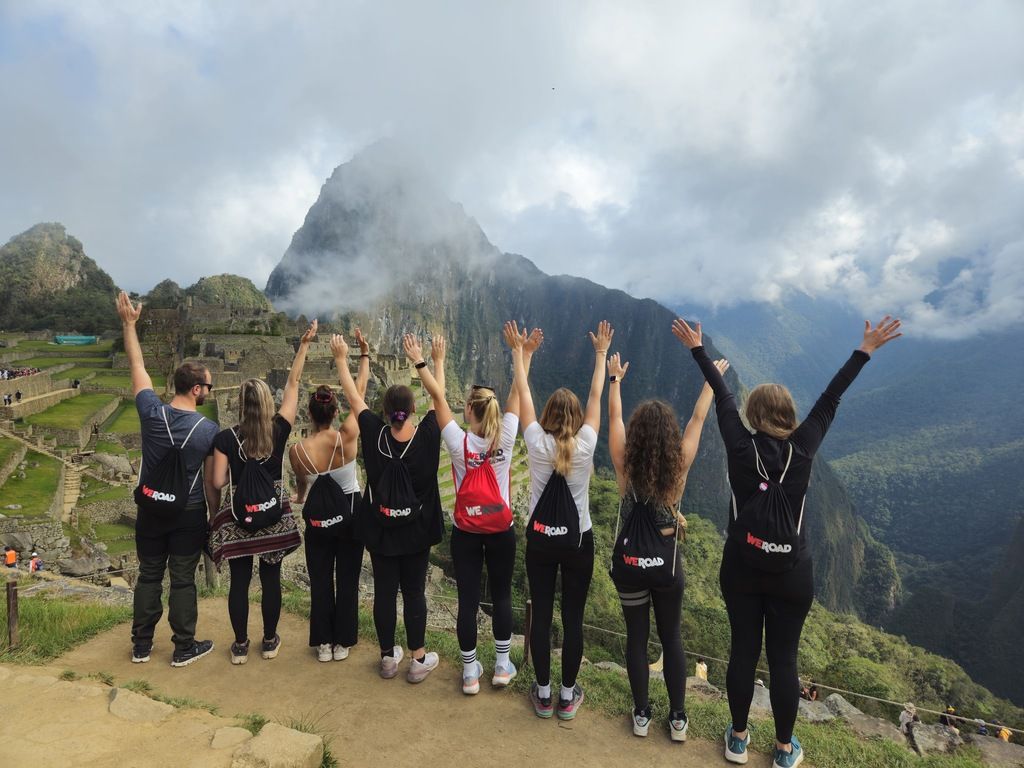 A group of travelers with WeRoad backpacks raising their hands at Machu Picchu, Peru, with mist-covered mountains in the background.