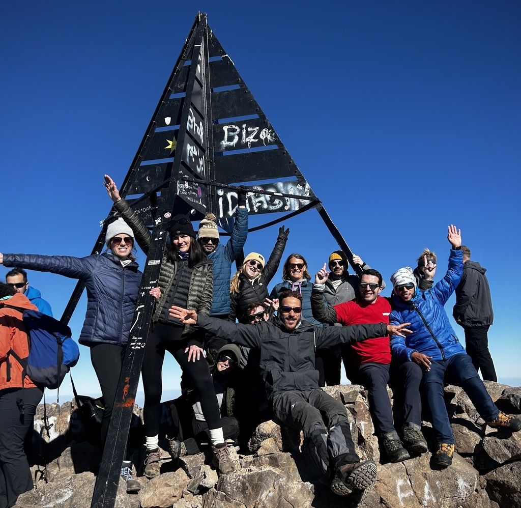 A group of hikers celebrating at the summit of Mount Toubkal, the highest peak in North Africa, posing next to the iconic metal pyramid marker.