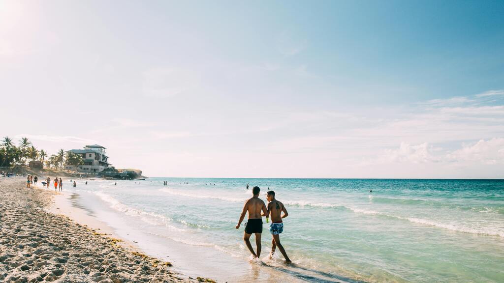 Two men walk along a sunny cuban beach with clear turquoise water.