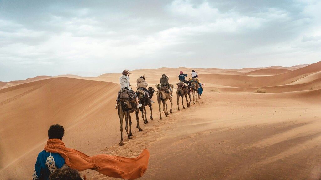 Group of travelers riding camels through the Sahara Desert under a cloudy sky