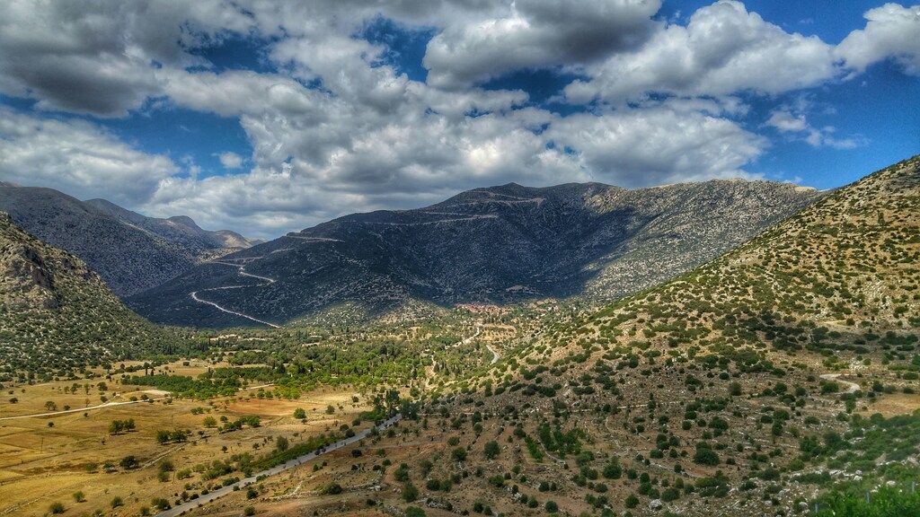 A winding road through a mountainous Greek landscape with green valleys and scattered trees under a dramatic cloudy sky.