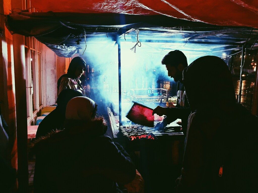 People gathered around a smoky barbecue grill at night, with one person fanning the flames.