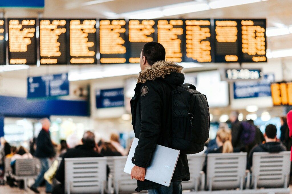 A man with a backpack holding a laptop looks up at a departure board in a busy airport terminal.