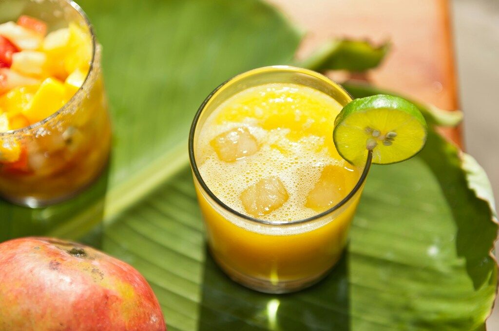 Frescos, a glass of yellow fruit juice with ice cubes and a lime slice on a green leaf, with another drink in the background.