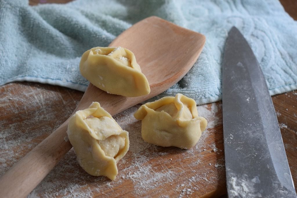 manti: three uncooked dumplings on a wooden board next to a wooden spoon and a knife, with flour dusted on the surface.