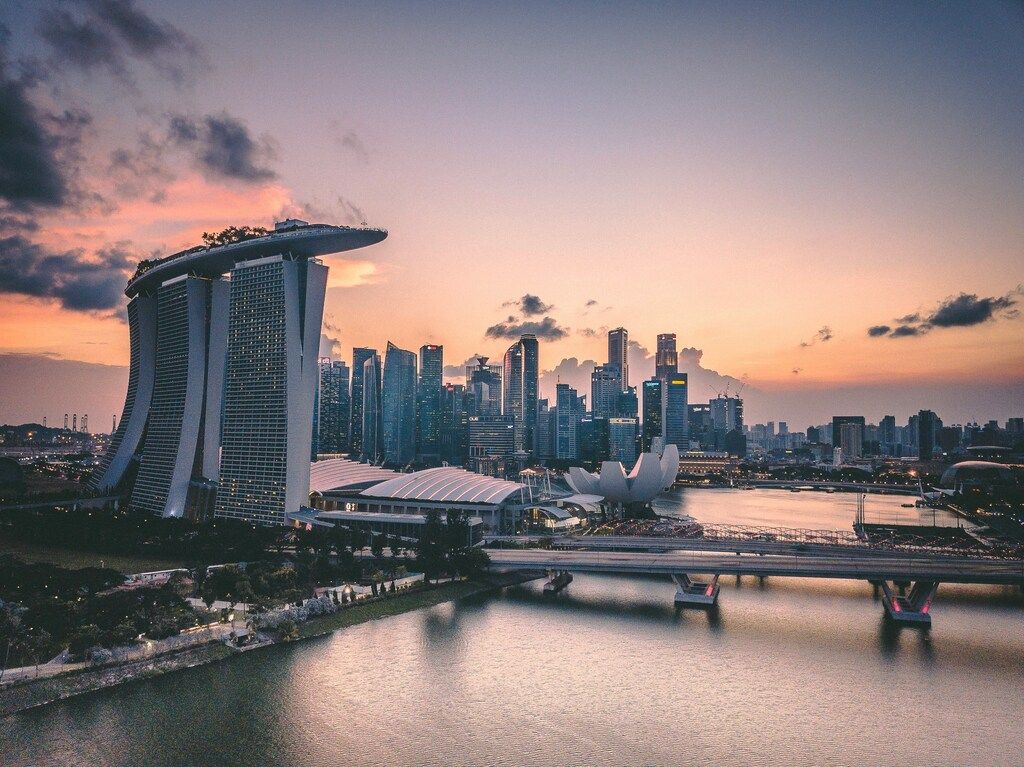 The modern skyline of Singapore at dusk, featuring the Marina Bay Sands hotel and other skyscrapers reflecting in the water, with a bridge spanning the bay.