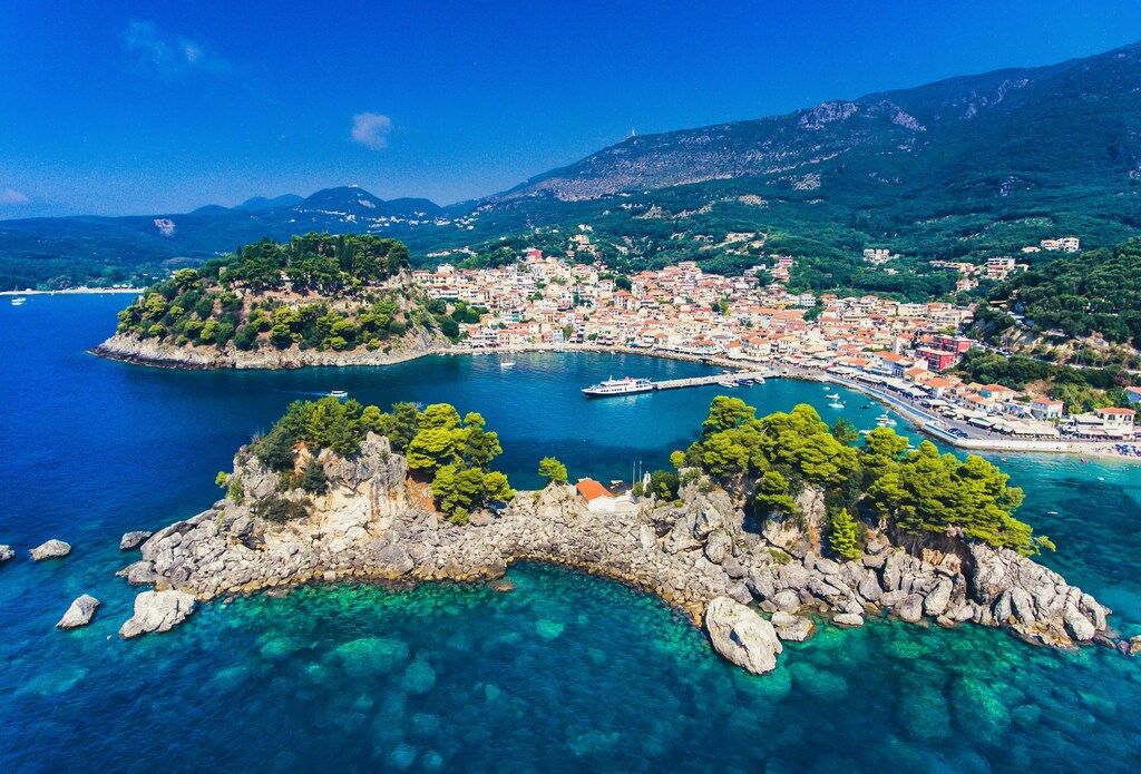 An aerial view of a picturesque Greek coastal town and bay with small islands, clear turquoise waters, and a mountainous backdrop.