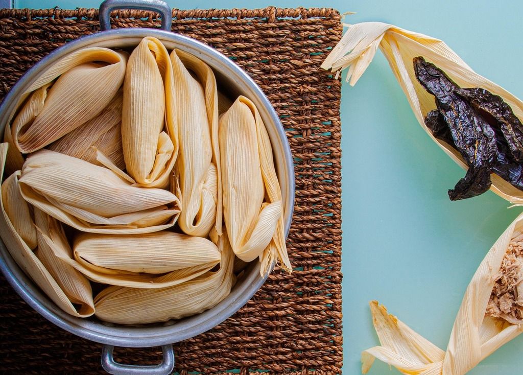 tamales: a metal pot filled with tamales wrapped in corn husks, next to additional husks with fillings.