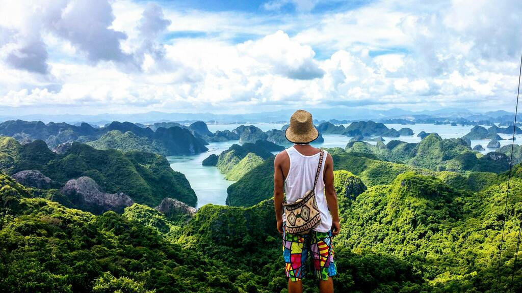 A person wearing a straw hat and colorful shorts stands on a lush green mountain overlooking a bay with numerous green limestone karsts under a cloudy sky.