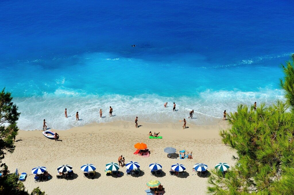 An aerial view of a vibrant blue ocean with waves crashing on a white sandy beach, dotted with beachgoers, umbrellas, and green trees in Greece.