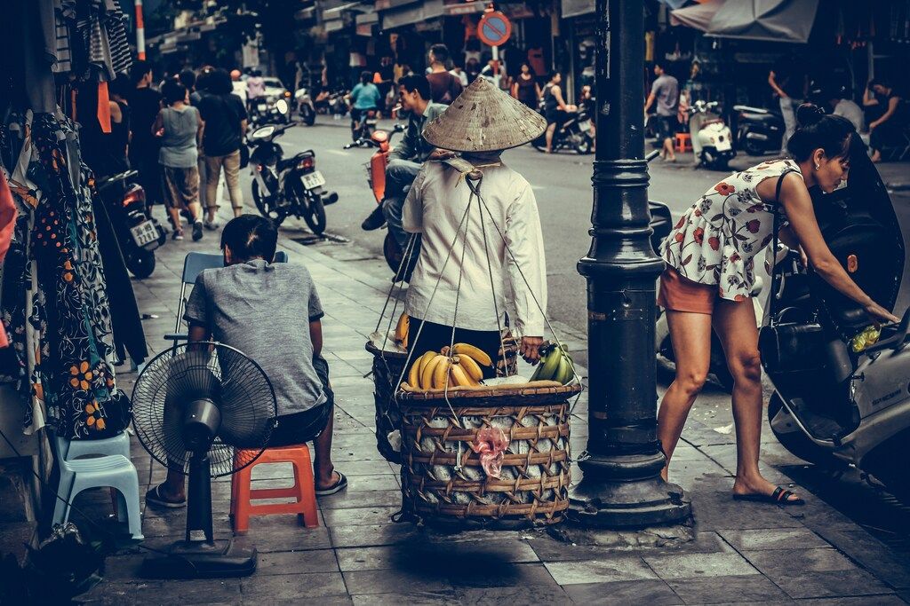Vietnamese woman carrying baskets of bananas on a yoke in a busy street market in Hanoi.