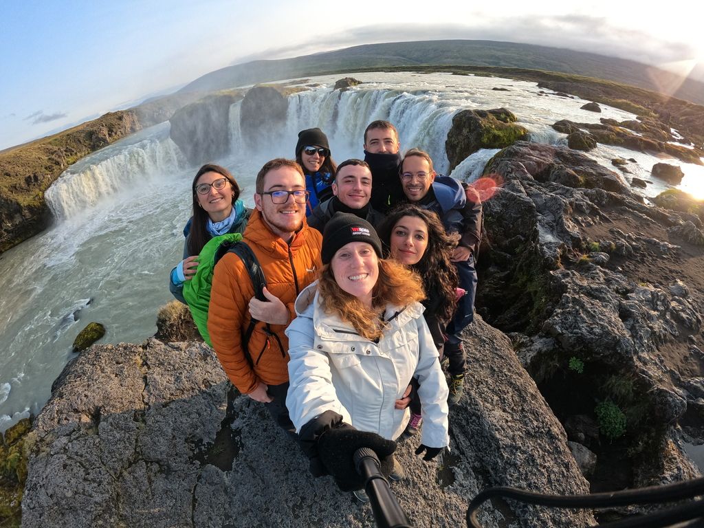 A group of happy WeRoad travelers taking a selfie together on the rocks at the top of a powerful waterfall in Iceland.