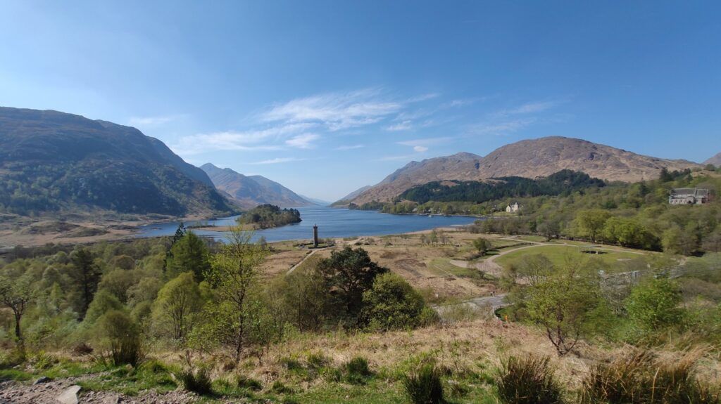A scenic view of the Scottish Highlands, showing a wide loch surrounded by mountains and a prominent monument by the water's edge.