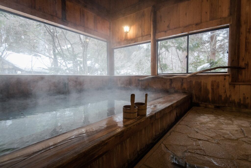 Steaming indoor wooden onsen with large windows showing a snowy landscape.