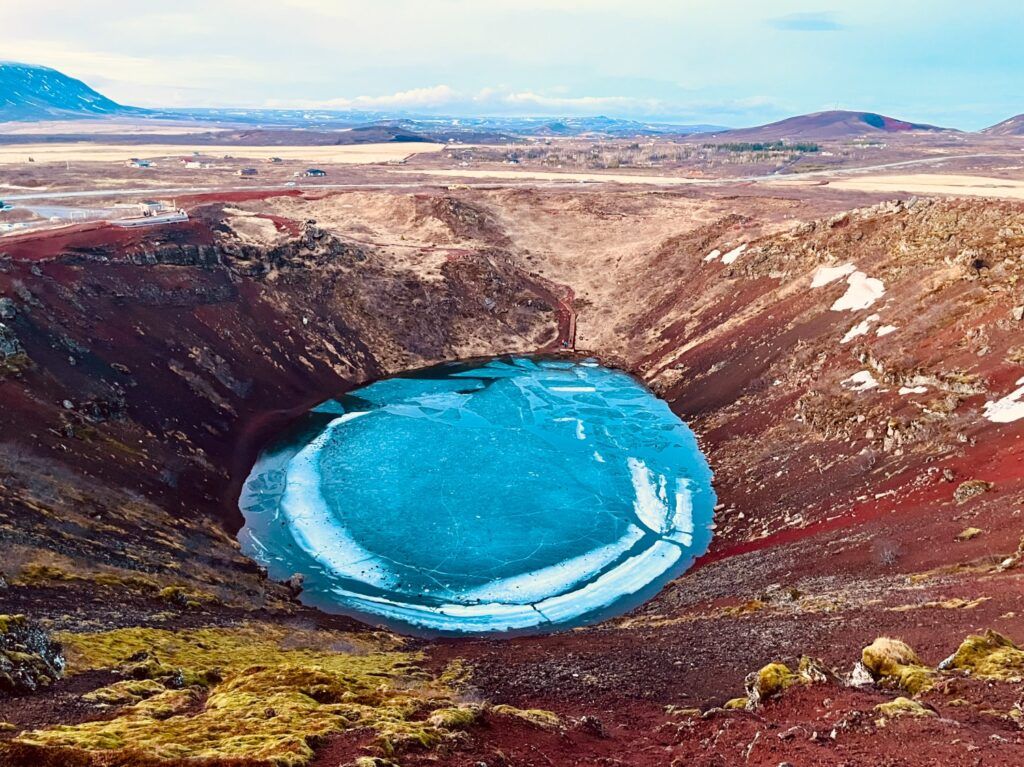 Aerial view of the striking Kerid volcanic crater with its bright blue, partially frozen lake surrounded by red earth.