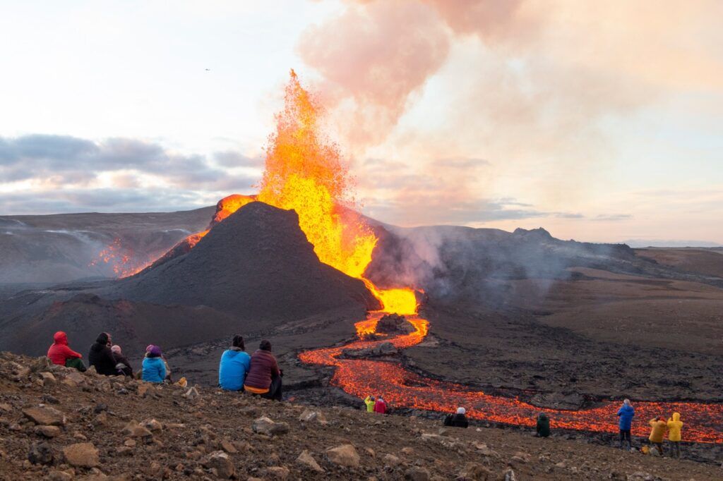 Spectators seated on a rocky hill watching a fiery volcanic eruption with lava flowing down the slope.