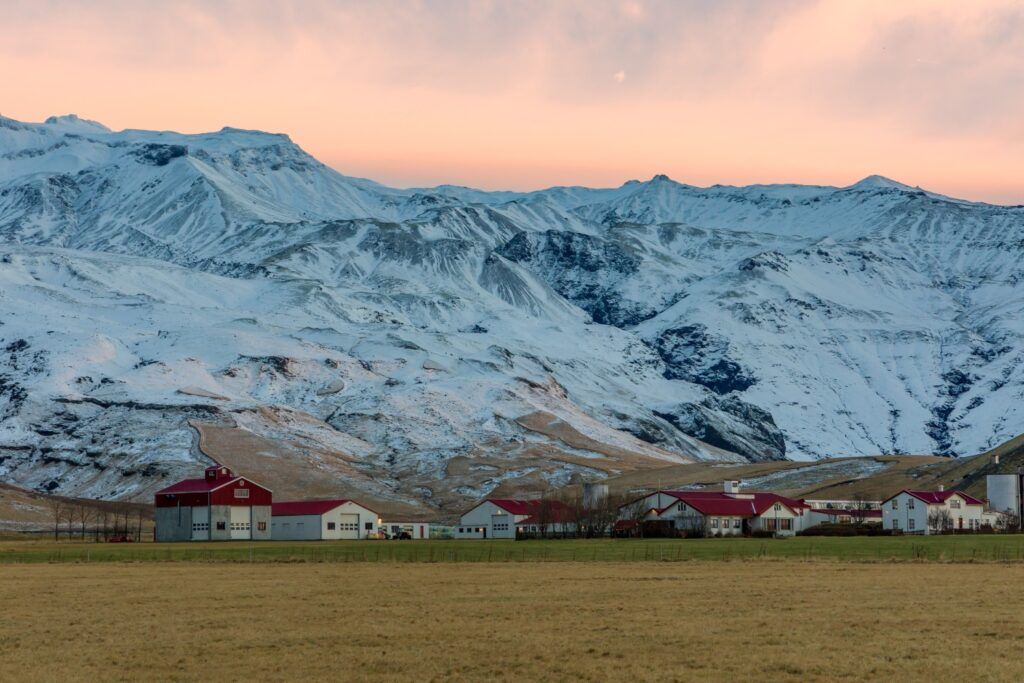 Red and white farm buildings nestled at the foot of the massive, snow-covered Eyjafjallajökull volcano in Iceland at sunset.