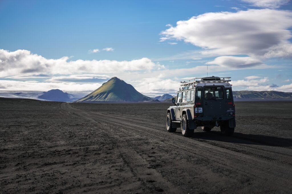 Land Rover driving on a black sand road toward a cone-shaped green mountain in Iceland.