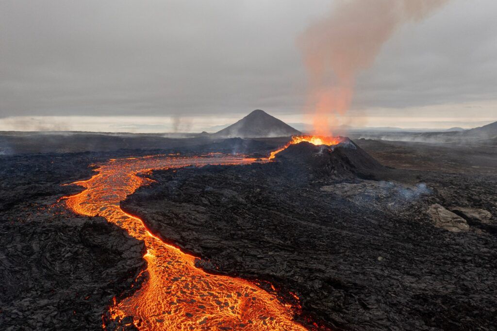 Aerial view of a river of bright, glowing orange lava flowing through black volcanic rock with steam rising.