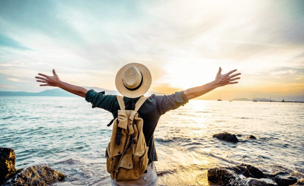 Solo traveler with a backpack and hat standing at the seashore with arms raised, celebrating discovery at sunset.