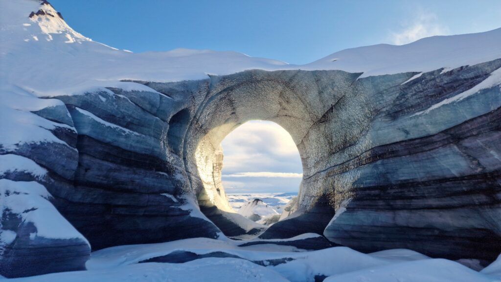 Natural ice arch or cave in a glacier with layered blue and white ice, framed by snow.