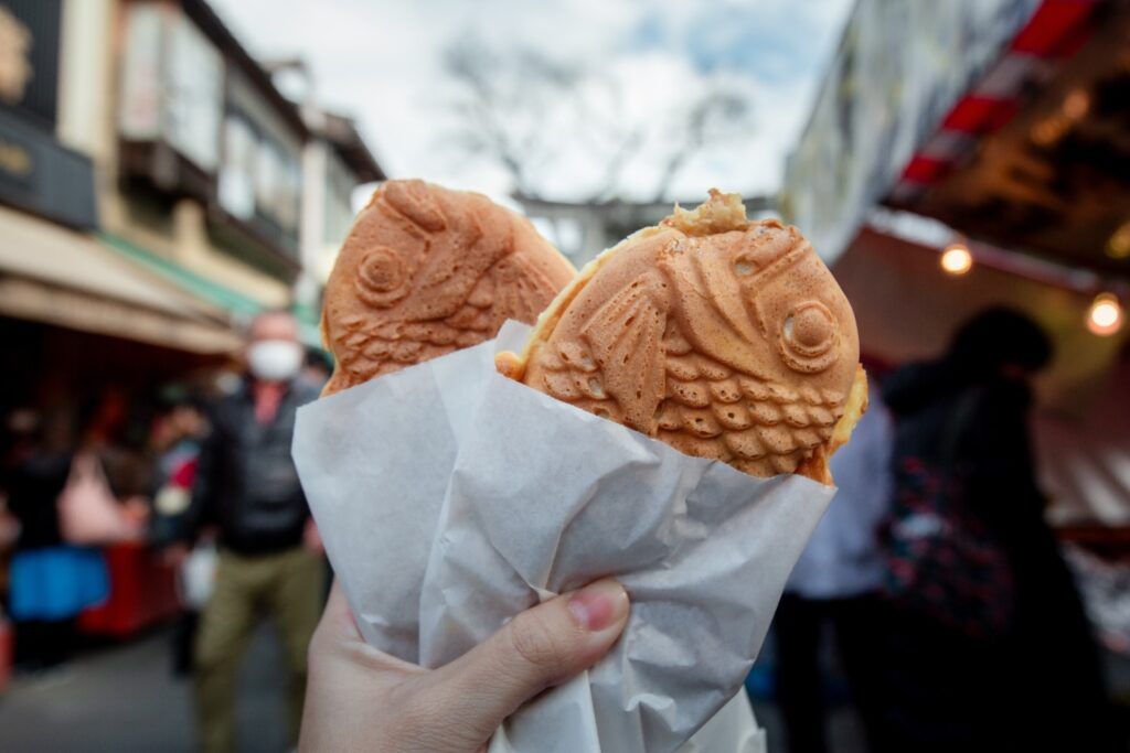 Hand holding two fish-shaped Taiyaki waffles filled with custard or red bean paste on a busy street.