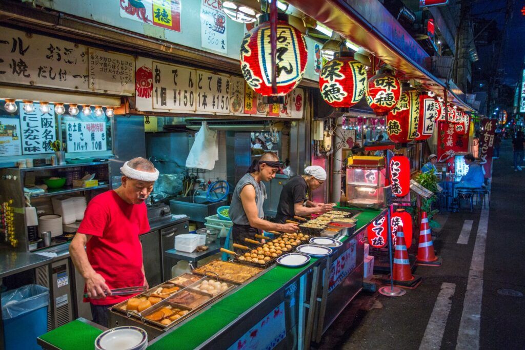 Vibrant Japanese night market scene with food stalls, red lanterns, and chefs cooking Takoyaki and Okonomiyaki